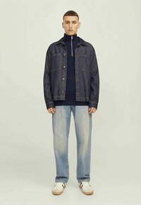 Mlad moški v temnem jeans jopiču, oblečenem preko mornarsko modre zip-up jopice, v svetlih zbledelih kavbojkah in belih supergah, stoji ob preprosti ozadju.