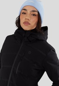 Czarna ocieplana kurtka z wysokim kołnierzem, z zamkniętą kieszenią i subtelną prążkowaną teksturą. W zestawie z jasnoniebieską czapką beanie.