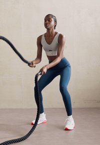 Fitnes atletka v sivi kratki majici in teal leginsih, ki uporablja črne bojna vrvi, oblečena v bele in roza teniske, na nevtralni podlagi.
