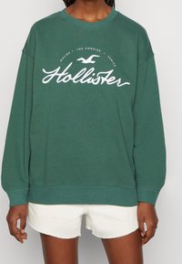 Vihreä collegepaita, jossa valkoinen "Hollister"-logo ja graafinen kuviointi, pyöreä pääntie ja resorit hihoissa. Yhdistetty valkoisiin shortseihin.