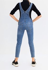 Blue denim overalls with a fitted shape, featuring open back design, tapered legs, and two back pockets. Worn with black sneakers.