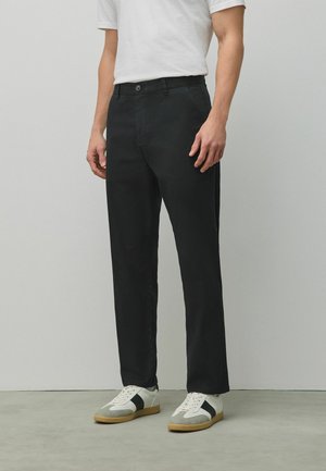 Man wearing black straight-leg pants, white t-shirt, and white sneakers with black stripe, standing on gray floor against light wall.