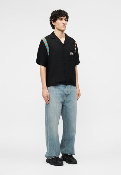 Άνδρας με σγουρά μαλλιά, φορώντας μαύρο κοντομάνικο πουκάμισο με πολύχρωμες ρίγες στους ώμους, ανοιχτόχρωμο τζιν και μαύρα αθλητικά παπούτσια, στέκεται ουδέτερα.