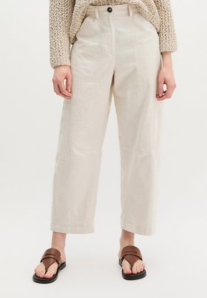 Beżowe szerokie spodnie z wysokim stanem i naszywanymi kieszeniami, w połączeniu z grubym beżowym swetrem oraz brązowymi skórzanymi sandałami z czarnymi podeszwami.