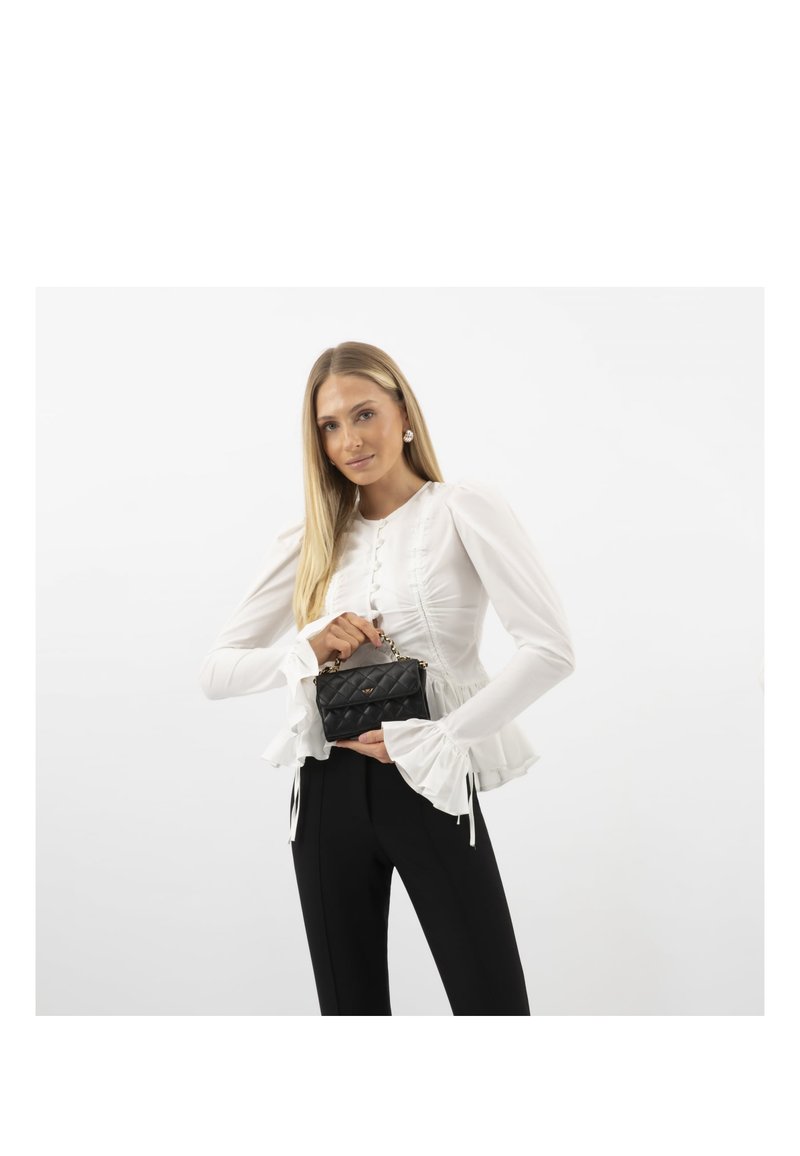 Femme en blouse blanche et pantalon noir tenant un petit sac à main noir matelassé avec une bandoulière chaîne sur fond blanc.