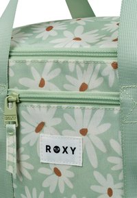 Groene tas met bloemenprint, versierd met witte madeliefjes en bruine centra. Heeft een ritsluiting en een "ROXY" label. Getextureerde stof.