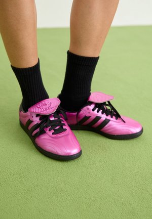 Benenden met zwarte sokken en felroze Adidas sneakers met zwarte strepen op een groen tapijt.