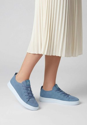 Ben in een witte geplooide rok wearing blauwe gebreide sneakers met witte zolen, staand op een lichtgrijze vloer.