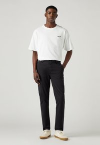 White cotton t-shirt with black logo, paired with black pants and beige sneakers. Simple design with relaxed fit and classic style.