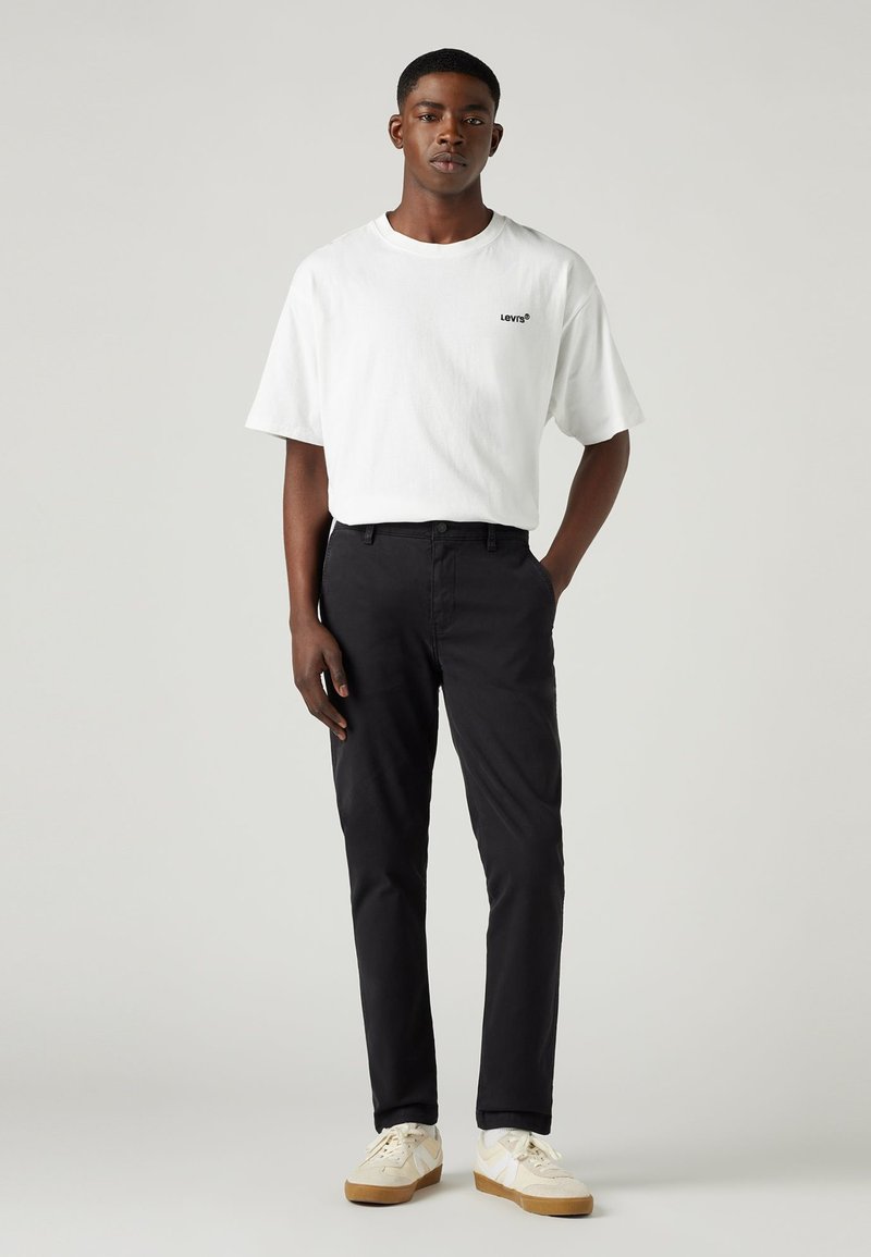 White cotton t-shirt with black logo, paired with black pants and beige sneakers. Simple design with relaxed fit and classic style.