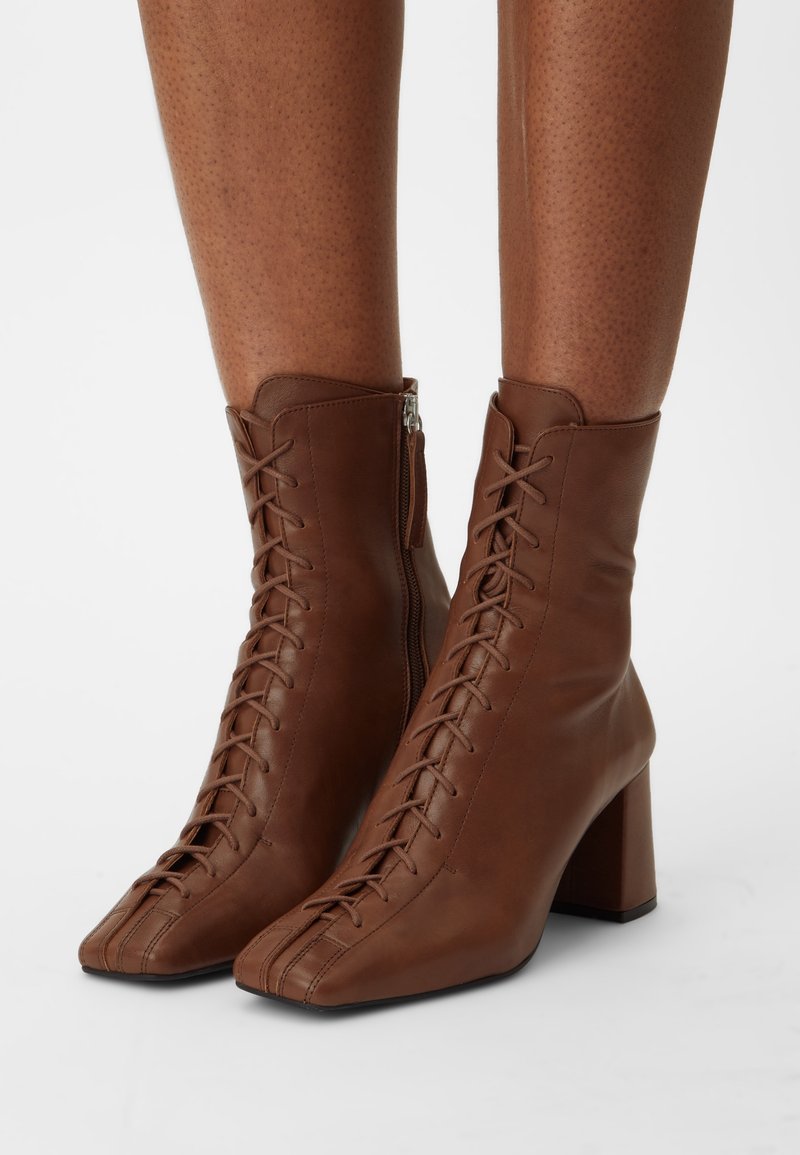 Bottes chelsea en cuir marron avec lacets à l'avant, fermeture éclair latérale, bout carré et talons épais de hauteur moyenne portées sur des jambes nues.