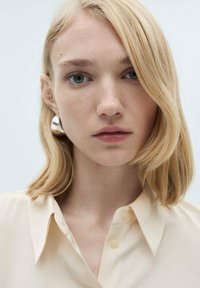 Blondes Haar mit sanften Wellen; trägt ein hellbeiges Hemd mit Kragen. Auffällige runde, metallische Ohrringe und ein neutraler Hintergrund.