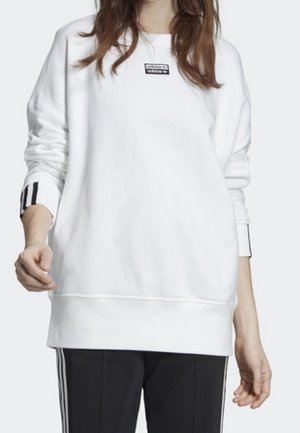 Femme portant un sweat-shirt blanc à manches longues avec un petit logo sur la poitrine et un pantalon noir avec des bandes blanches sur les côtés.