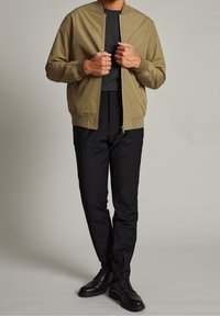 Chaqueta bomber color caqui con cierre de cremallera, puños de canalé y una camiseta negra de cuello redondo debajo, combinada con pantalones negros y zapatos negros.