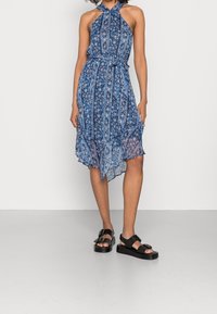 Blå blommig klänning med halterneck och bälte i midjan; asymmetrisk nederkant, lättviktig tyg, kombinerad med kraftiga svarta sandaler.