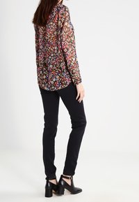 Blommönstrad, genomskinlig blus i flera färger, tillsammans med åtsittande svarta byxor och svarta klackade ankelstövlar. Sedd från baksidan.