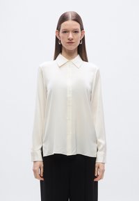 Long-sleeve cream blouse in soft fabric, featuring a classic collar, button front, and smooth texture. Paired with black trousers.