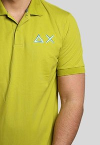 Polo verde con colletto, caratterizzato da un logo geometrico azzurro chiaro sul petto. Realizzato in tessuto liscio, con maniche corte e una forma aderente.