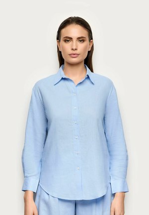 Donna in piedi rivolta in avanti che indossa una camicia azzurro chiaro con bottoni e maniche lunghe e pantaloni coordinati su uno sfondo semplice.