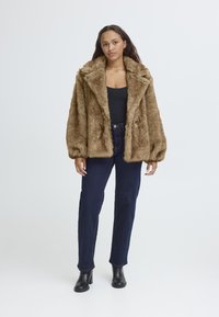 Giacca in finta pelliccia marrone con ampio colletto e grandi rever, vestibilità comoda, superficie testurizzata, abbinata a jeans scuri a vita alta e stivali neri.