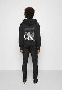 Svart hoodie med en stor vit "CK" logotyp på baksidan, i kombination med svarta cargobyxor. Materialet verkar mjukt; designen är avslappnad och funktionell.