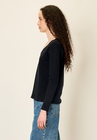 Ženska z dolgimi kodrastimi lasmi, oblečena v črno majico s dolgimi rokavi in modre jeans hlače, stoji postrani na enobarvni svetli podlagi.