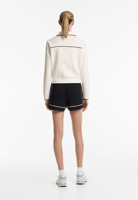 White sweatshirt with black piping, paired with black shorts featuring white trim. Model is wearing light grey sneakers and white socks.
