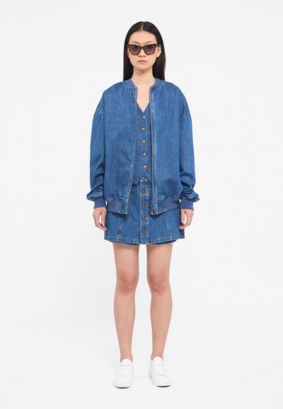 Denim jacket with a zip front and ribbed cuffs, paired with a denim skirt featuring a button closure. Both are light blue. White sneakers worn.