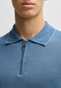 Blå zip-up polo tröja med krage och kontrasterande sömnadsdetaljer. Tillverkad av ett strukturerat stickat tyg. Närbild av halslinningen.