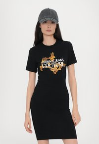Crna majica haljina s oblikovanim siluetom i grafičkim printom s natpisom "VERSACE JEANS COUTURE" u zlatnoj i bijeloj boji. Nosena s sivom kapom.