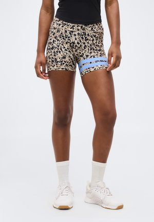 Žena v športových šortkách s leopardím vzorom a dvoma modrými pruhmi na jednom stehienku, bielych ponožkách, bielych teniskách a čiernom topu.