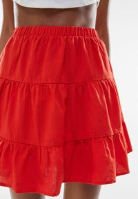 En kvinna i en ljus röd, flerdelad kjol med resårmidja och en vit kortärmad topp mot en neutral vit bakgrund.