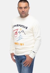 Maglione color crema con la scritta "Courmayeur" e grafiche di sci in blu, rosso e arancione. Realizzato in tessuto morbido con polsini e fondo a coste.