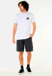 T-shirt bianca con un motivo circolare nero, abbinata a pantaloni cargo scuri fino al ginocchio e scarpe da ginnastica nere con suola bianca.