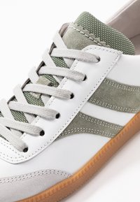 White sneaker with green suede accents, light gray laces, textured green tongue, and gum rubber sole, shown in close-up detail.