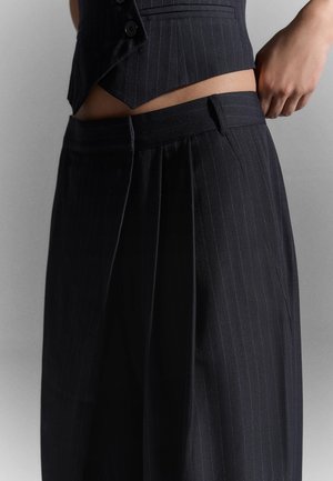 Henkilö, joka pukeutuu tummaan liituraitaiseen pukuun, jossa on liivi ja leveälahkeiset housut, säätää vyötärönauhaa yksiväristä vaaleaa taustaa vasten.