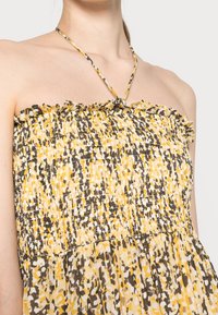 Šaty bez ramínek s nařaseným živůtkem, zdobené abstraktním vzorem v žluté, hnědé a bílé barvě. Volánkové okraje a tenké zkřížené ramínka.
