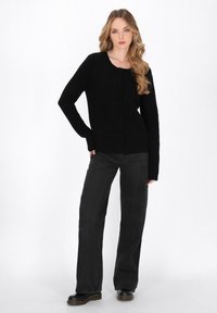 Donna con lunghi capelli mossi, che indossa un cardigan nero con bottoni, jeans neri a gamba larga e stivali neri, in piedi con una mano in vita.