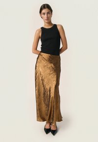 Top casual nero a coste abbinato a una lunga gonna bronzo lucida con una finitura testurizzata. La modella indossa tacchi a punta neri.