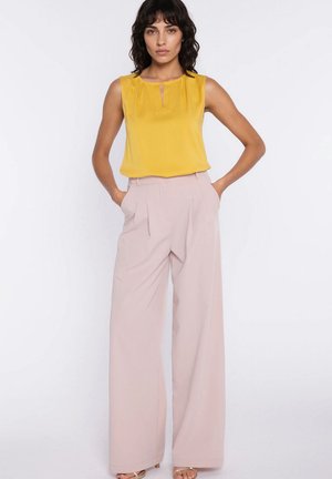 Kobieta o ciemnych włosach, ubrana w bezrękawnik w kolorze musztardowym, wsunięty do wysokich, szerokich beżowych spodni, stojąca z rękami w kieszeniach.