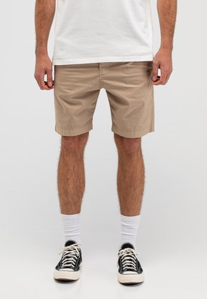 Mann trägt beige Shorts, weiße Socken, schwarze Turnschuhe und ein weißes T-Shirt, steht vor einem schlichten, hellen Hintergrund.