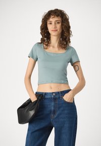 Camiseta corta de color azul claro con mangas cortas, escote cuadrado, combinada con pantalones vaqueros de mezclilla azules de talle alto y un bolso tipo pouch negro.