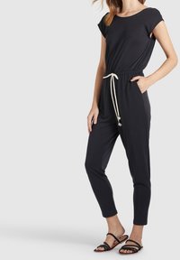 Zwarte jumpsuit met korte mouwen, trekkoord in de taille, zijzakken en taps toelopende broekspijpen. Draag het met zwarte bandjesandalen. Glad stoffen textuur.