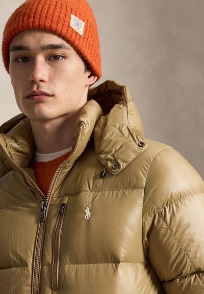 Jeune homme portant un bonnet tricoté orange et une veste matelassée beige avec un col montant, une fermeture éclair visible et un logo sur la poitrine.