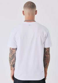 Camiseta blanca de manga corta con un acento azul cerca del escote, hecha de una tela suave. La vista posterior muestra brazos tatuados.