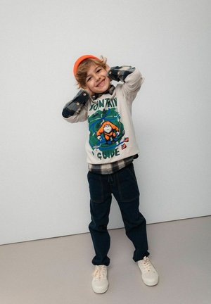 Uśmiechnięte dziecko w pomarańczowej czapce, graficznej bluzie z napisem "Przewodnik Górski", w kratkowanej koszuli, ciemnych spodniach i białych sneakersach, pozujące z rękami za głową.