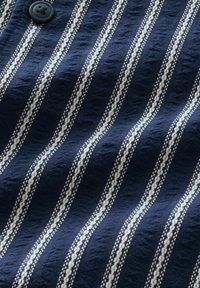 Námořnická modrá tkanina s vertikálními bílými pruhy, s texturovaným vzorem. Obsahuje detail v podobě černého knoflíku. Měkký, lehký materiál.