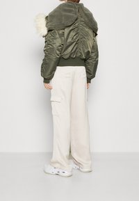 Chaqueta bomber verde oliva con un cuello texturizado forrado de piel; material suave y brillante; combinada con pantalones cargo anchos en beige y zapatillas blancas.