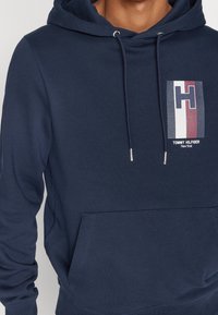 Sudadera azul marino con un bolsillo tipo canguro, cordones y un logotipo rectangular que presenta letras en negrita y rayas en rojo, blanco y azul.