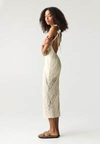 Ein cremefarbiges Kleid aus strukturiertem, gemustertem Stoff, mit offenem Rücken und gebundenen Trägern, kombiniert mit braunen Sandalen mit dekorativen Schnallen.